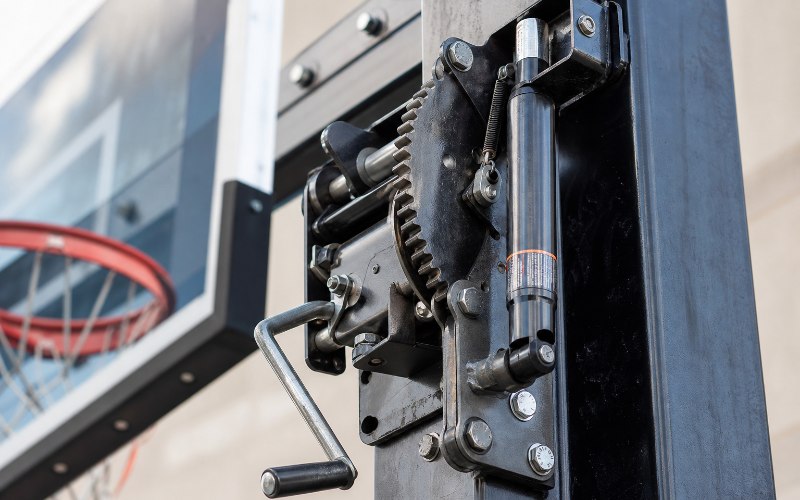 Close-up view of a hydraulic or crank-based height adjustment mechanism on an in-ground basketball hoop pole.